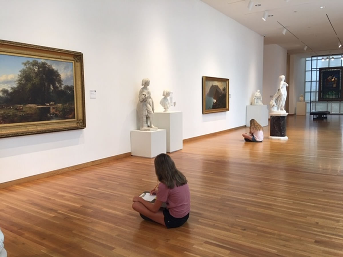 two socially distanced girls sit on the floor in the gallery and draw the landscapes they see on the walls.