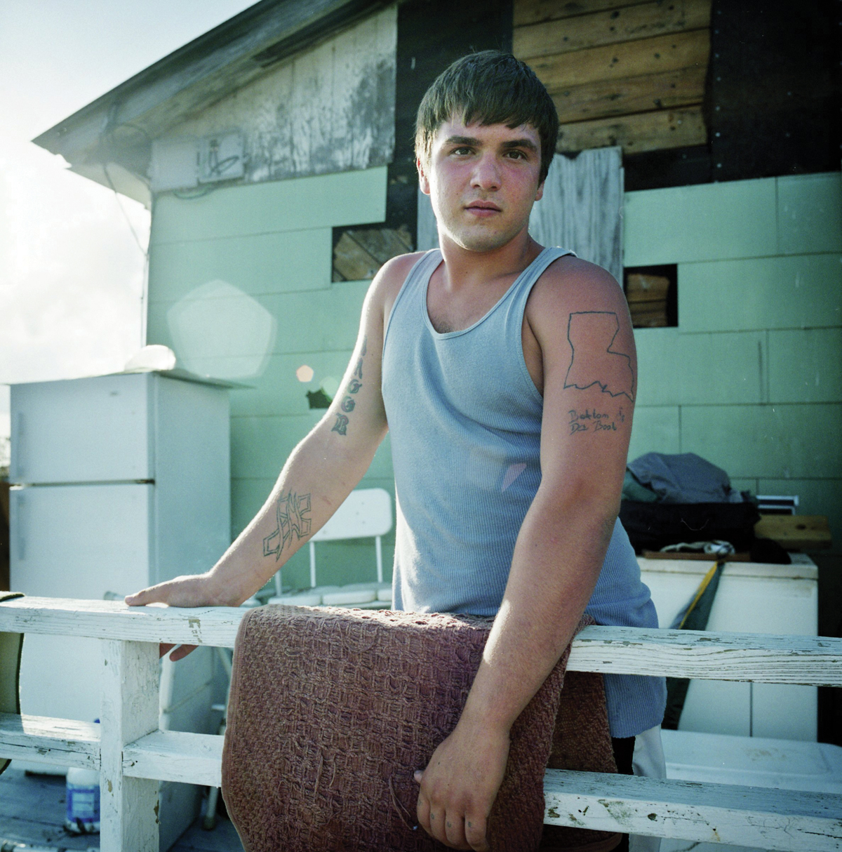 A man in a blue tank top stands at the railing of a porch; tattoos on his arm include the state of Louisiana and the phrase "Bottom of the Boot."