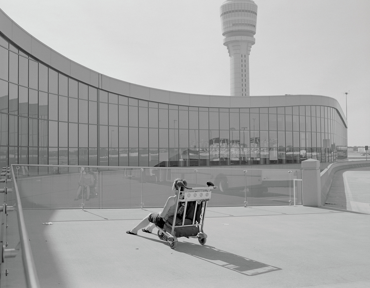 A woman sits on a luggage trolley facing a glass railing across from the curved, glass-fronted airport building.