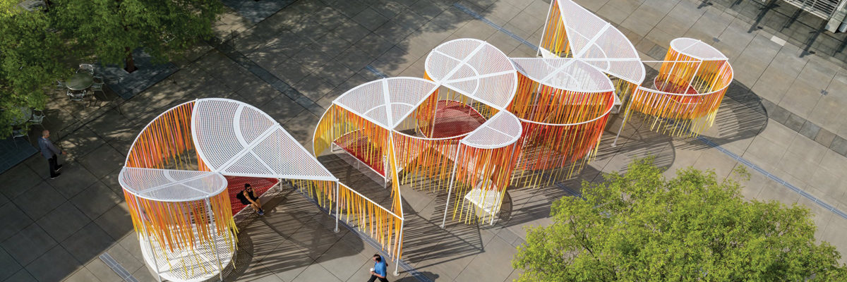 Top of art installation in the Piazza from above; white mesh interlocking shapes with red, yellow, and orange strips hang beneath them.
