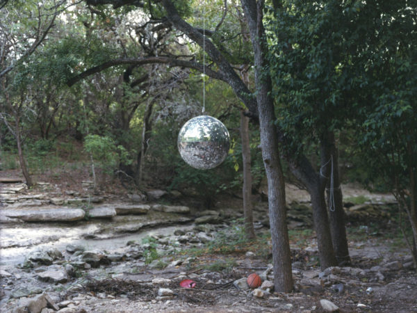 A disco ball hangs from a branch in the woods near a dry creek bed; a basketball and red frisbee lay on the ground nearby.