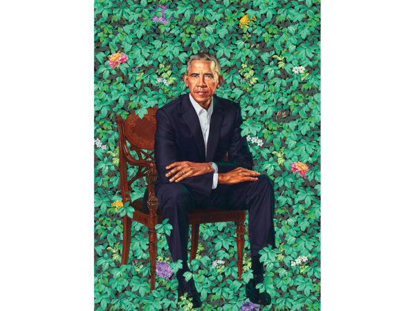 Former President Barack Obama, wearing a dark suit, sits in a wooden chair against a full background of ivy scattered with multi-colored blooms.
