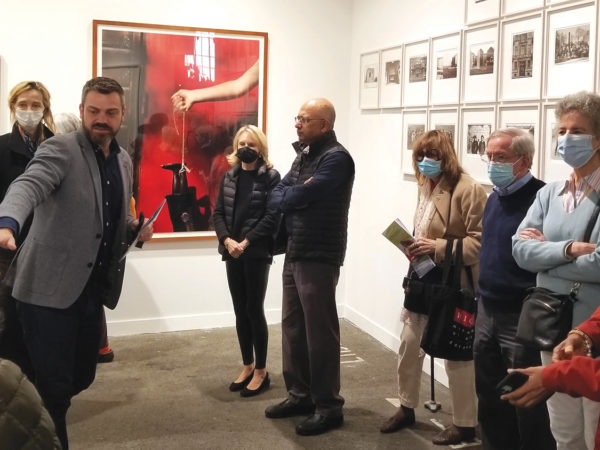 A group of people stand in a small gallery of framed photos while a curator points to the wall next to him.