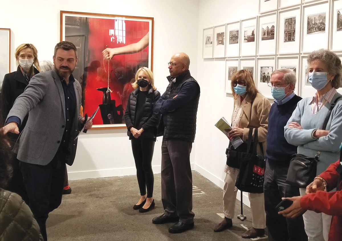 A group of people stand in front in a small gallery hung with framed photos while a curator points to the wall next to him.