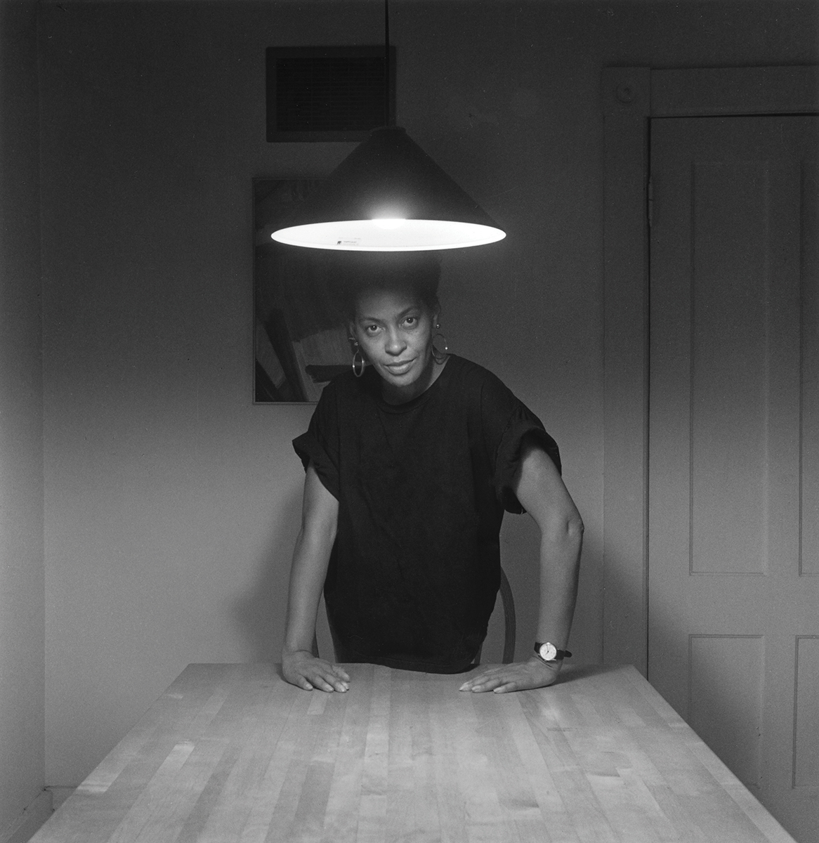 A woman stands with her hands on a kitchen table beneath a hanging lamp, looking directly at the viewer.