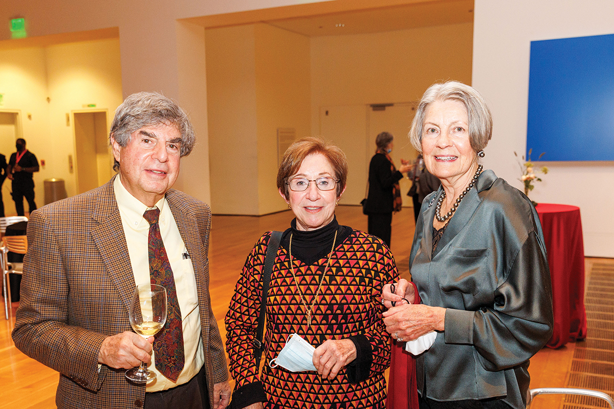 Three people stand together in the lobby, smiling toward the camera; one of them holds a wine glass.