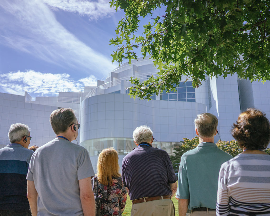 A group of people with their backs to the camera and each wearing earphones stand outside under a tree and look toward a large white-tiled building.