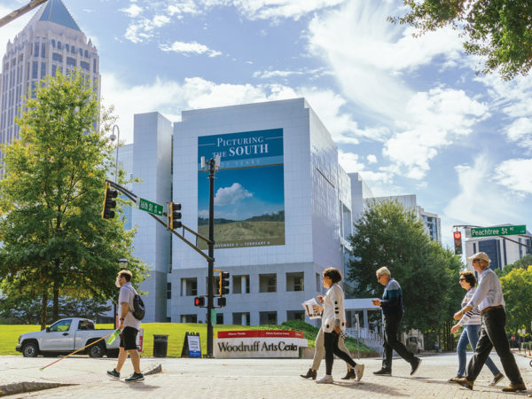 A group of people cross an intersection with the building banner for Picturing the South: 25 Years hanging on the High Museum of Art in the background.