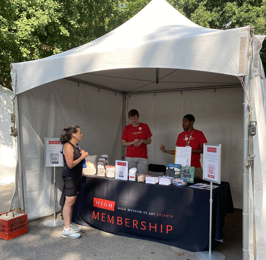 Two volunteers wearing red t-shirts talk to a visitor from behind a High Museum membership table stacked with brochures.