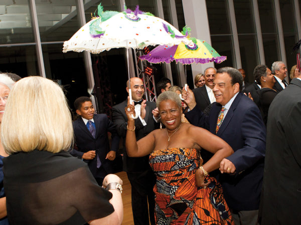 Two people hold Mardi-Gras-themed umbrellas aloft while dancing through a crowd of people in the museum lobby.