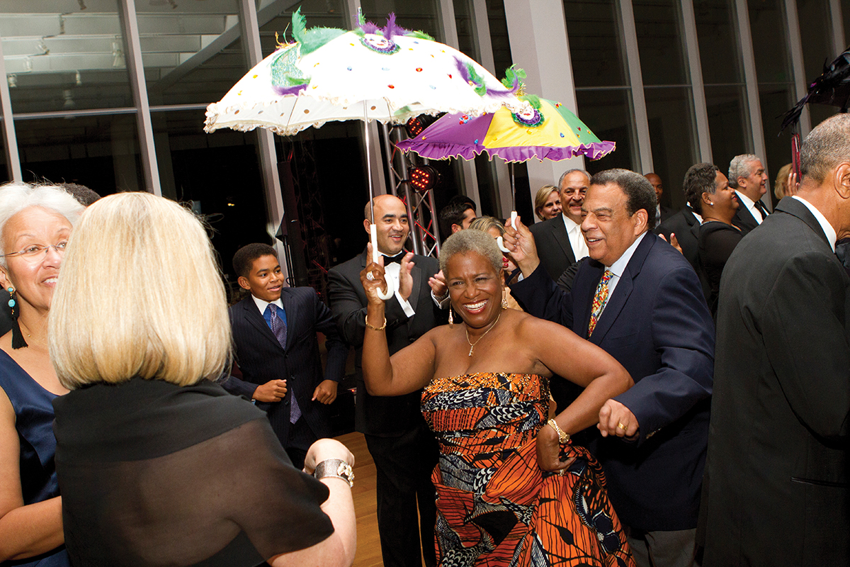 Two people hold Mardi-Gras-themed umbrellas aloft while dancing through a crowd of people in the museum lobby.