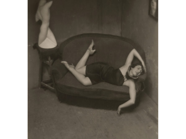 A woman in a short black dress reclines on a loveseat, her legs and arms twisted into a pose. A statue of a nude torso stands on a small table to the left of the loveseat in a similar pose.