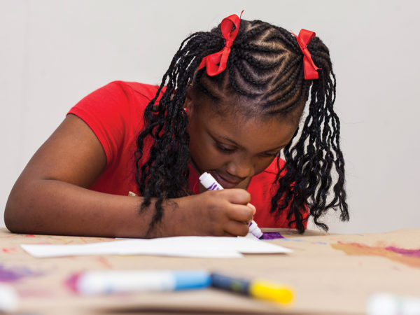 A girl with red ribbons in her braids draws on a table-sized piece of paper with a purple marker.