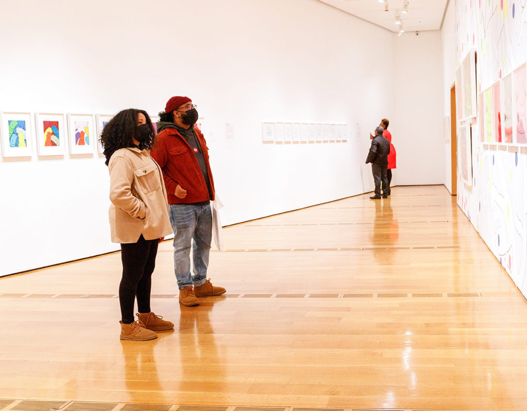 Two visitors stand in the center a gallery to view prints on a wall while two people in the background stand close to read a wall label.