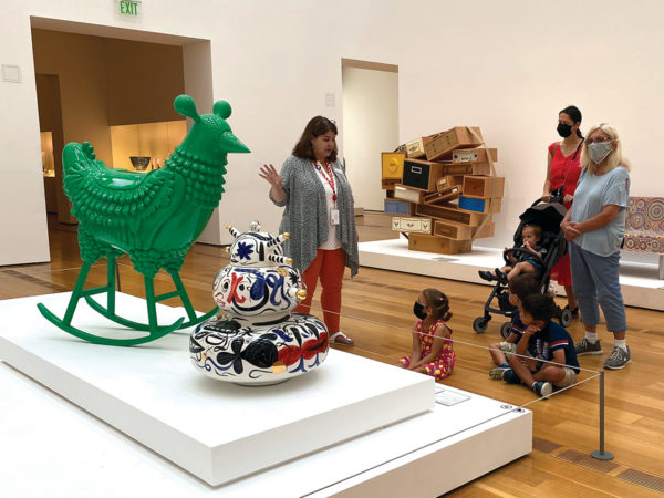 A staff member gestures toward two colorful sculptures on a pedestal while talking to visitors and their children in a gallery.