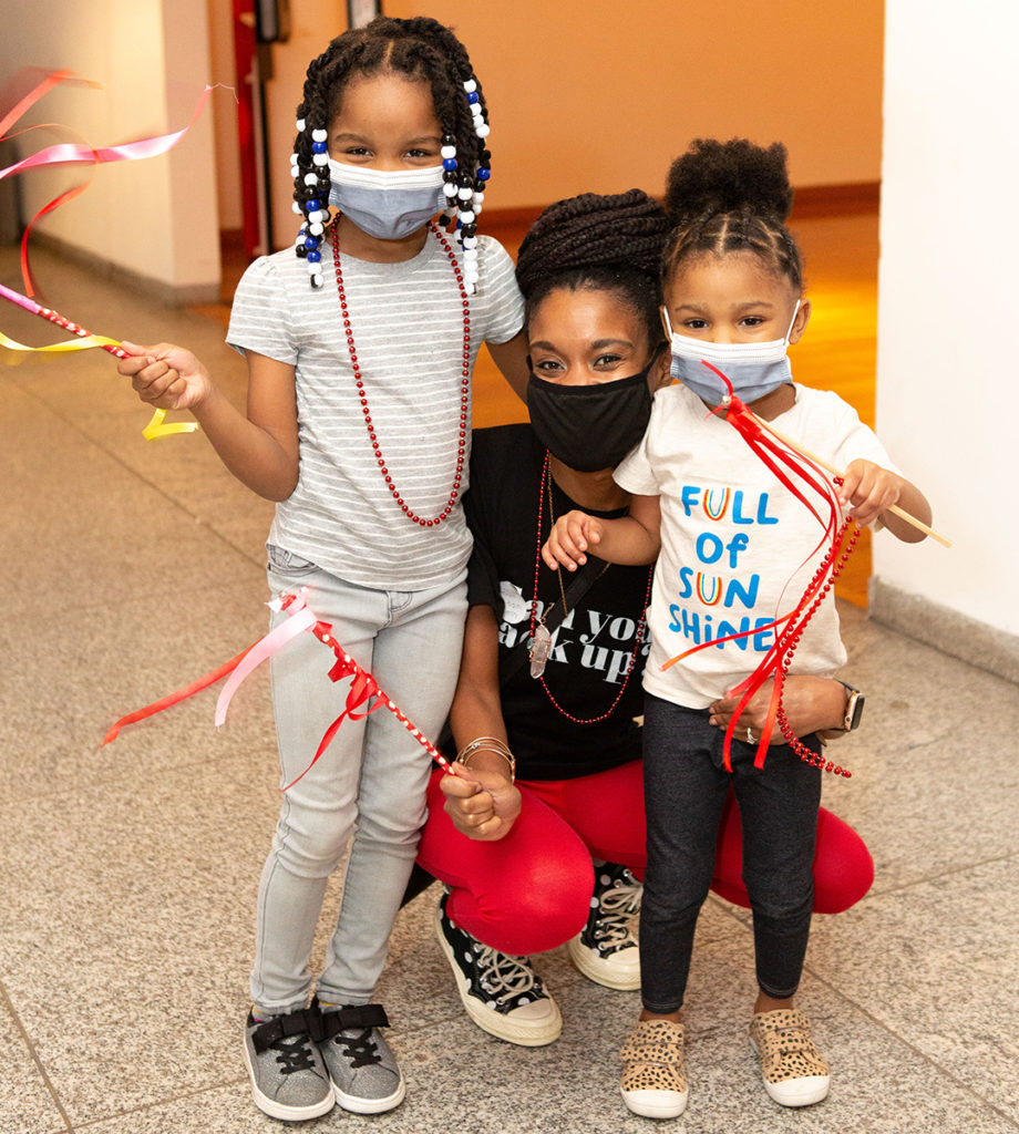 A visitor with two young children pose for the camera while waving sticks decorated with ribbons and beads.