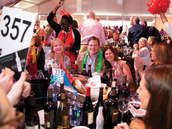 A group of people, several of them smiling at the camera, sit around a large table covered with wine bottles and food. Beyond them are more tables full of people within a large decorated tent.