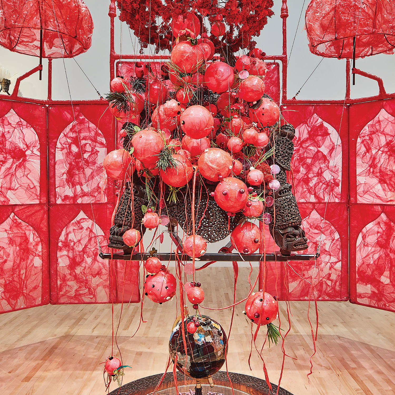 Installation piece with translucent, decorative red barrier surrounding a bundle of decorative red balls around a carved wooden sculpture above a globe in the center of the floor.