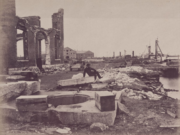 A man sits atop a slab of stone looking toward docked boats, the ruins of a large brick building behind him.