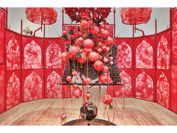 Installation piece with translucent, decorative red barrier surrounding a bundle of decorative red balls around a carved wooden sculpture above a globe in the center of the floor.