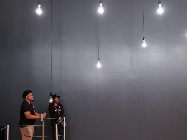 Two visitors in Rafael Lozano-Hemmer's Pulse Room. One person holds the metal sensors to have their pulse recorded, while the two people look at the lightbulbs hanging from the ceiling.