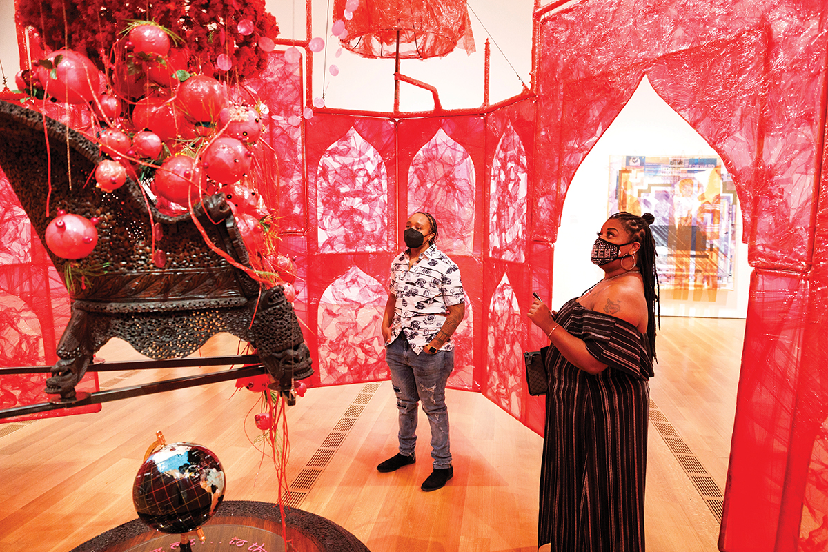 Two visitors stand inside a decorative red, palace-shaped structure and look at a black chair hanging from the center of the structure and filled with red balls.
