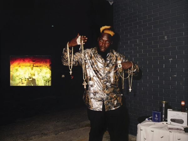 A photograph of a Black man holding up jewelry while a photograph behind him shows a Black man in a field holding up a garden tool.