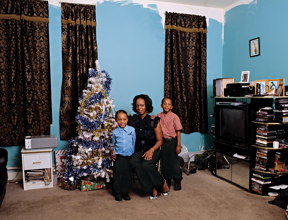 A woman and her two kids pose in their home next to a decorative blue, white, and gold tree with presents underneath. The blue walls in the background are only partially painted.