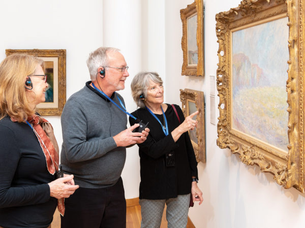 Three people listening to an audio tour view a painting in a gallery.