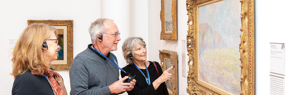 Three people listening to an audio tour view a painting in a gallery.