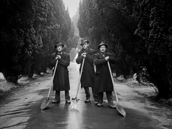 Black and white photograph of three men in overcoats pose in a tree-lined road, each leaning on a shovel.
