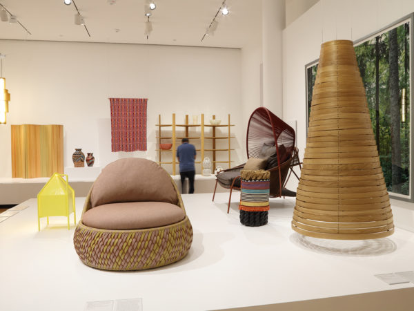 A visitor looks a set of shelves behind a platform with chairs and a large wooden cylinder in the gallery of the Stephen Burks exhibition.
