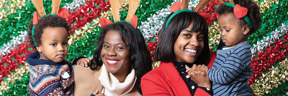 Two women, each holding a small child pose for the camera in front of a backdrop of red, green, gold, and silver bows.