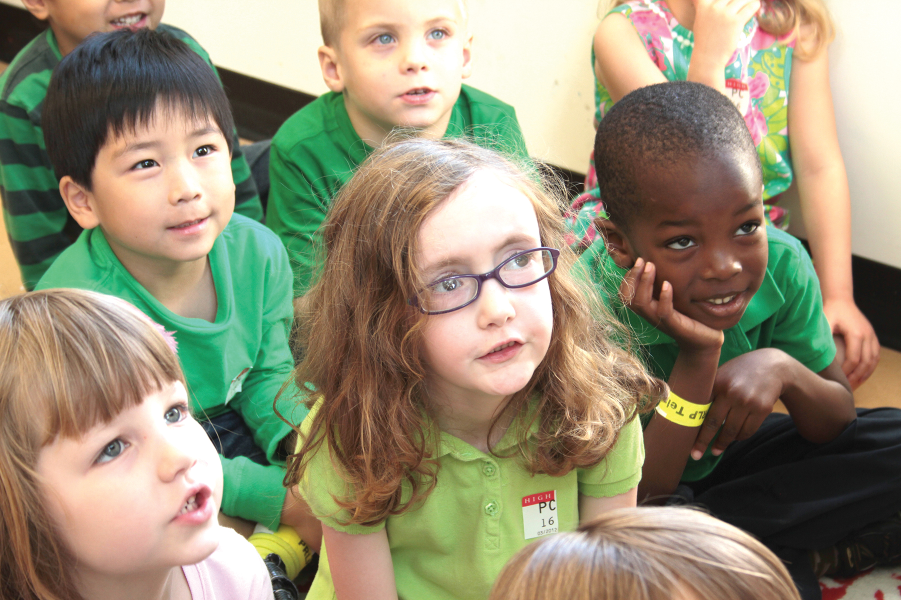 A group of children sit in a group looking towards something out of view.